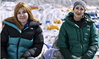 Two women in winter jackets pose outdoors in a snowy area, smiling at the camera.