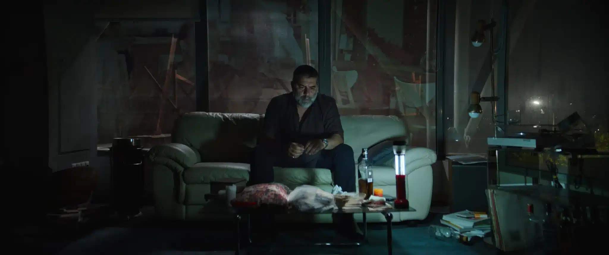 Man with gray beard sits on a beige sofa in a dimly lit, cluttered living room, looking down at his hands,